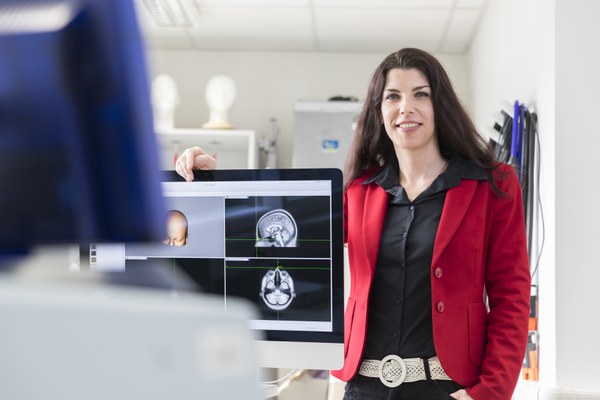 Frau Daun lehnt mit einem Arm über einem Bildschirm und lächelt freundlich in die Kamera. Auf dem Bildschirm sind MRT-Scans eines menschlichen Gehirns sowie eine Rekonstruktion eines Kopfes zu sehen.